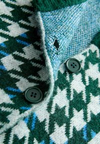 Groene en witte gebreide cardigan met een patroon, voorzien van donkergroene knopen en een close-up van een knoopsgat op een gestructureerde stof.