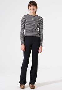Zwart-wit gestreepte top met lange mouwen, gecombineerd met zwarte flared broek en gemusterde bruine schoenen. Effen achtergrond, model staat lachend.