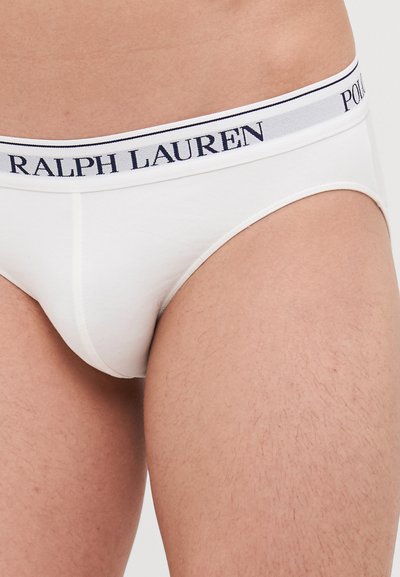 Bele moške hlačke v beli barvi z logom na pasu, na katerem sta napisana "RALPH LAUREN" in "POL" v mornarsko modri barvi. Narejene so iz gladkega materiala z oprijetim krojem.