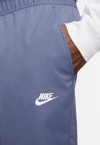 Sinisiä urheiluhousuja sileästä kankaasta, joissa on sivutasku ja valkoinen Nike-logo vasemmassa alakulmassa.