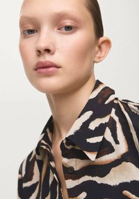 Srajca s potiskom tigrovih črt v krem, črni in rjavi barvi. Iz gladkega blaga, z klasičnim ovratnikom in izpostavljenimi gumbi.