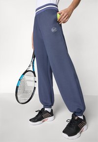 Person som holder et svart og blått tennisracket og en grønn tennisball, iført marineblå joggebukser og svarte sportsko med hvite såler.