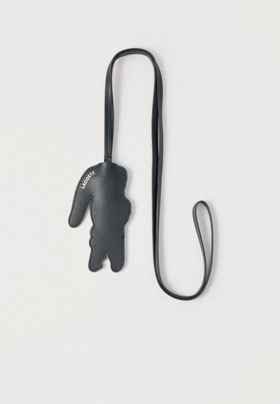 Porte-clés en cuir noir en forme de crocodile avec la marque "Lacoste" et une longue sangle fine, sur un fond blanc uni.