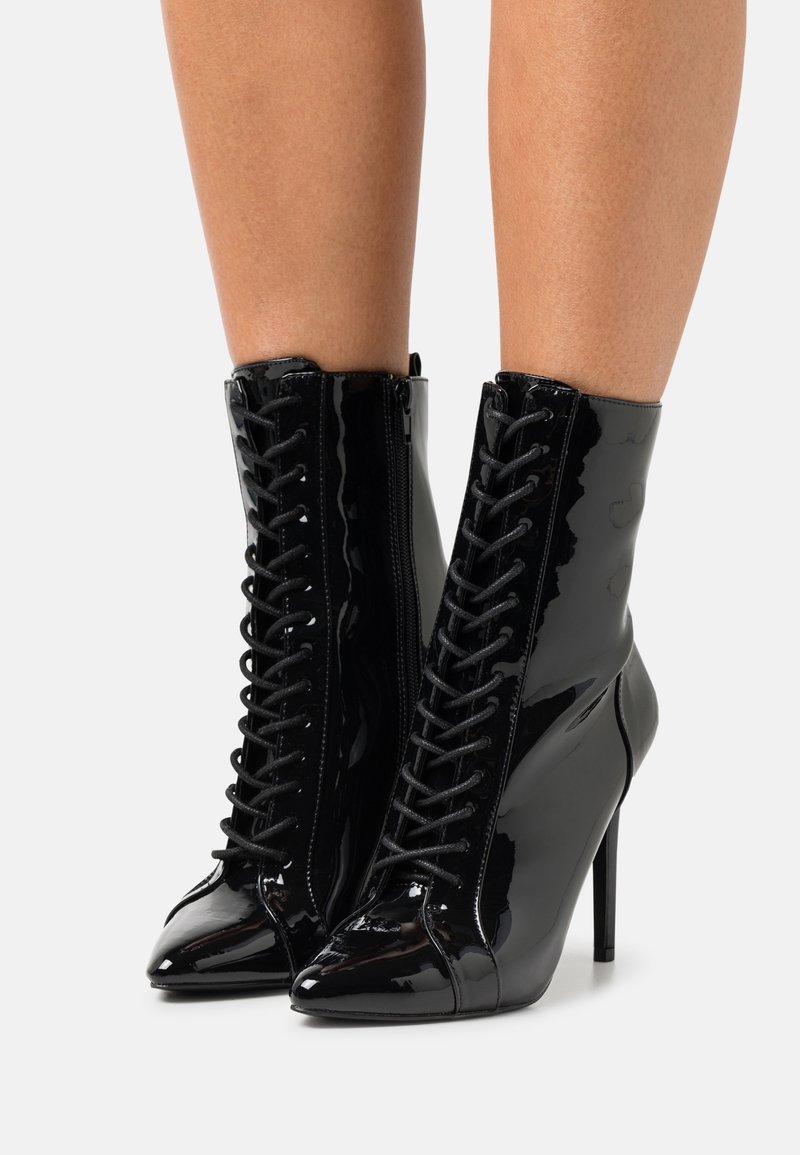 Bottines en cuir verni noir avec bout pointu, talon stiletto haut, laçage à l'avant et fermeture éclair à l'arrière.