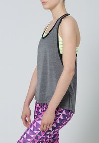 Canotta grigia senza maniche realizzata in tessuto leggero, con profilo nero, indossata sopra un reggiseno sportivo a fantasia neon, abbinata a leggings geometrici rosa.