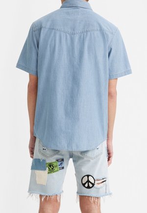 Homme portant une chemise bleu clair à manches courtes et un short en denim clair avec des patchs et des déchirures, vu de dos.