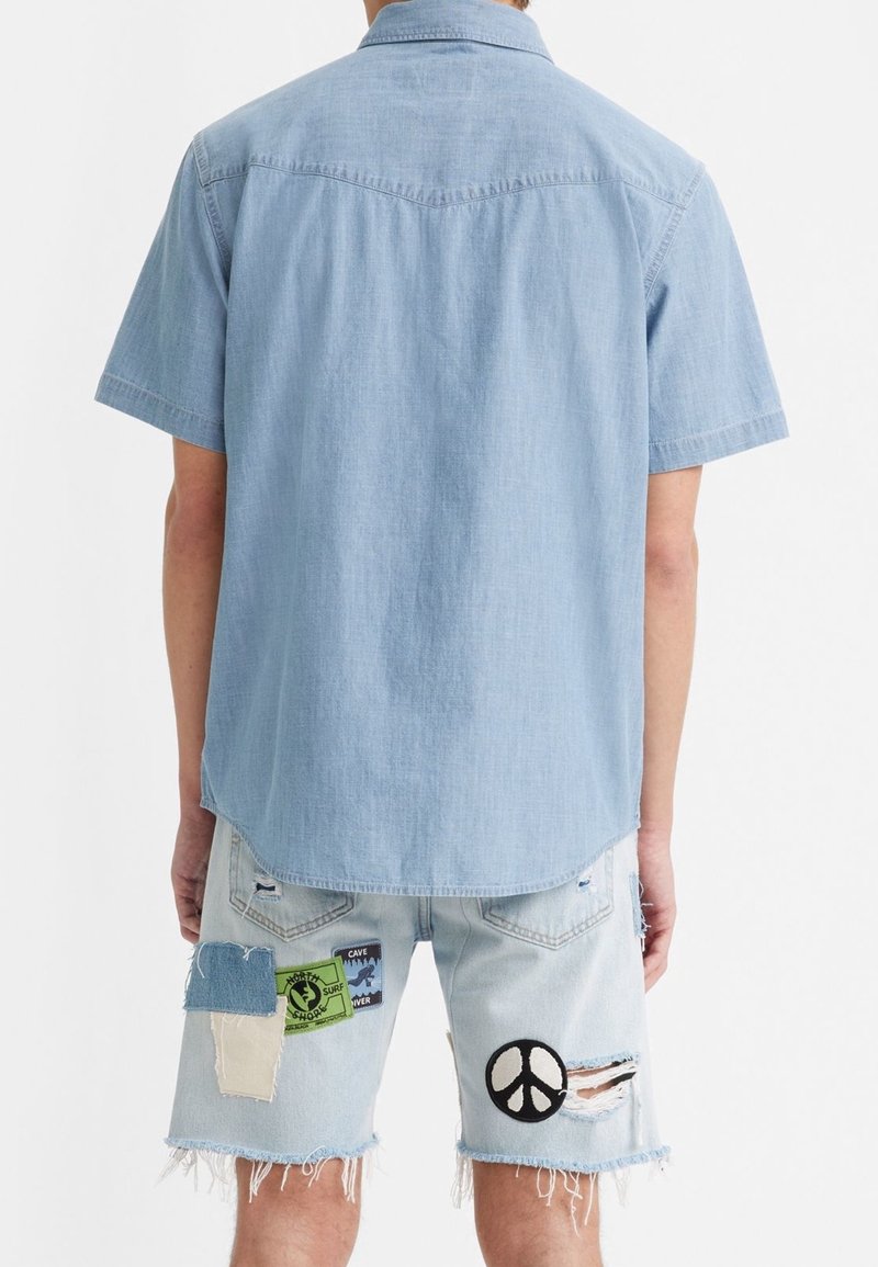 Homme portant une chemise bleu clair à manches courtes et un short en denim clair avec des patchs et des déchirures, vu de dos.