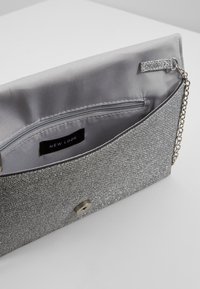 Pochette argentée pailletée ouverte laissant apparaître la doublure en tissu gris, une poche intérieure zippée avec une étiquette New Look, et une petite chaîne attachée à l’intérieur.