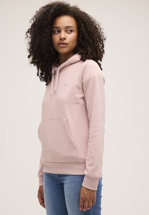 Mujer con pelo rizado que lleva una sudadera con capucha FILA de color rosa claro y vaqueros azules, mirando hacia un lado con un fondo liso.