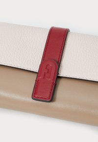 Läderplånbok med en texturerad krämfärgad utsida, slät beige bas, röd stängningsrem och en rund logotypdetalj.