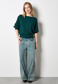 Junge Frau in einem strukturierten türkisfarbenen Pullover und weit geschnittenen, hellblauen Jeans, die mit einer Hand in der Tasche vor einer schlichten weißen Wand steht.