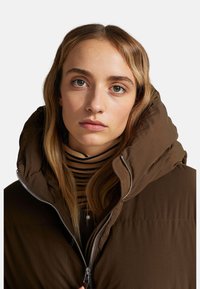 Hnědá péřovka s vysokým límcem, která má zipové zapínání a prošívanou strukturu. Pod ní je vidět černé a béžové pruhované tričko.