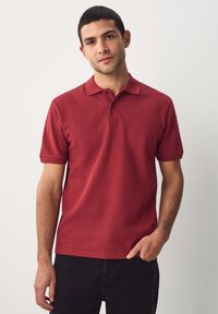 Polo rouge en tissu texturé, manches courtes et patte de boutonnage à trois boutons. Associé à un pantalon noir sur un fond neutre.