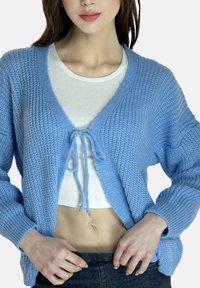 Hellblauer gestrickter Cardigan mit V-Ausschnitt, gebundenem Vorderteil und weiten Ärmeln, getragen über einem weißen Crop-Top und kombiniert mit dunklen Jeans.