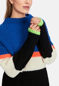Gestrickter Pullover mit blauen, schwarzen, weißen und orangefarbenen Streifen sowie grünen Rippbündchen. Die Textur ist dick und gemütlich.