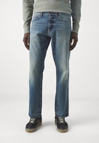 Man i urblekta blå jeans, grå långärmad skjorta och svarta sneakers med beige sulor som står mot en vit bakgrund.