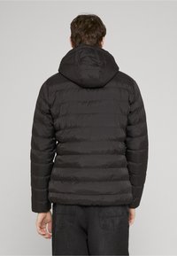 Doudoune noire à capuche, avec un design matelassé et une texture lisse et brillante. Fermeture éclair à l'avant et poignets élastiques.