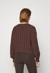 Persona con un suéter de punto trenzado de color marrón oscuro y pantalones a juego, visto desde la espalda contra un fondo claro y simple.