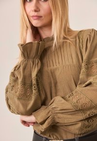 Mujer con cabello rubio largo vestida con una blusa marrón suelta con detalles de encaje en las mangas abullonadas y una tela texturizada, posando con una mano cerca del cuello.