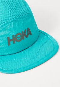 Turkusowa czapka o teksturowanym wykończeniu, z szarym logo "HOKA" i wyprofilawanym daszkiem. Szwy wzdłuż krawędzi podkreślają design.