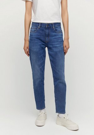 Persoon draagt mid-rise blauwe spijkerbroek, witte sokken, witte sneakers en een eenvoudig wit T-shirt met korte mouwen, staand tegen een effen achtergrond.