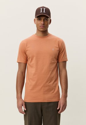 Giovane uomo che indossa un cappellino marrone con logo bianco "II", camicia a maniche corte color salmone con piccolo emblema "II" e pantaloni scuri, in posizione neutra.