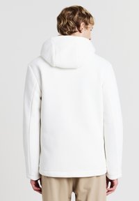 Weißer Kapuzenjacke mit geripptem Textur, lockerer Passform und leicht abgerundetem Saum. Die Ärmel sind lang und gerade ohne sichtbare Verschlüsse.