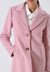 Donna che indossa un cappotto di lana rosa pastello con rever e due bottoni, abbinato a una camicia bianca con colletto e fermaglio dorato al collo.