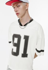 Hvid oversized jersey med sorte tal 91 og striber på ærmerne. Har et V-udskæring design, sølv kædede halskæde og en kasket.