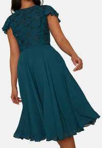 Teal klänning med korta volangärmar, blommig spetsliv och en flödande kjol som faller under knäet. Slät tyg med en mjuk textur.