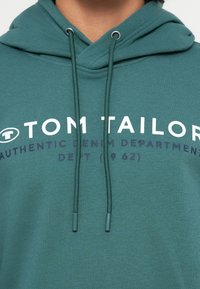 Felpa con cappuccio color teal, in misto cotone, presenta un cappuccio con cordino, stampa logo frontale "TOM TAILOR" e "AUTHENTIC DENIM DEPARTMENT" in bianco e viola.