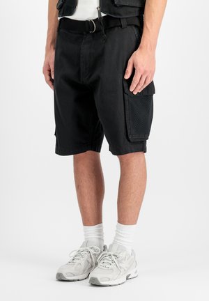 Mand iført sorte cargobukser med netlommer, hvide sneakers og hvide crew-sokker mod en enkel lys baggrund.