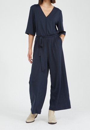 Laivansinis jumpsuit V-niskalla, lyhyillä hihoilla ja vyötäröltä vyötettynä. Siinä on leveät lahkeet ja pehmeä, kevytrakenne.