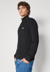 Maglione a coste nere con colletto alto e chiusura con zip, con un piccolo logo multicolore sul petto. Indossato con jeans blu.