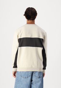 Sudadera beige claro con una franja horizontal negra, cuello redondo acanalado y puños elásticos. Textura suave, corte casual y colores contrastantes.