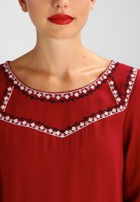 Rode blouse met een ronde halslijn, versierd met witte en zwarte bloemenborduursels langs de hals. Soepele stof en een comfortabele pasvorm.