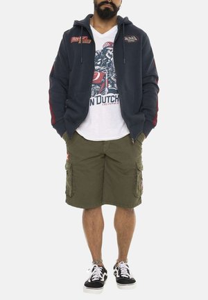 Sweat à capuche zippé navy avec des accents rouges, t-shirt graphique blanc avec un motif de crâne, shorts cargo olive et baskets noires.