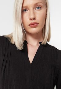 Czarna bluzka teksturowana z dekoltem w kształcie litery V i plisowanym wzorem. Zbliżenie pokazuje szczegóły materiału i akcenty stebnowania. Brak widocznych elementów metalowych.