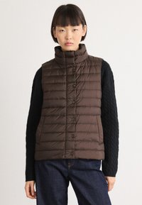Ungdom som har på seg en mørkebrun quillet ermeløs vest over en svart kabelstrikket genser og mørkeblå jeans, stående mot en enkel bakgrunn.