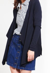 Cardigan blu scuro sopra una camicia a righe con colletto. Abbinato a una gonna di jeans con chiusura frontale a bottoni. Tessuto dalla texture liscia.