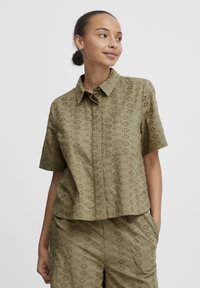 Short-sleeved, olive green shirt with a floral eyelet pattern, featuring a button-down collar and a chest pocket. Casual fit design.