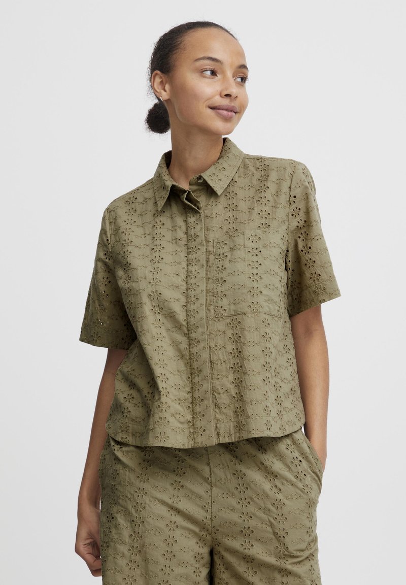 Short-sleeved, olive green shirt with a floral eyelet pattern, featuring a button-down collar and a chest pocket. Casual fit design.
