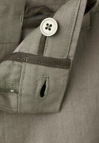 Makroaufnahme von khakifarbenem Stoff mit einem genähten Knopfloch und einem angenähten, beigefarbenen Vierlochknopf an einem gefalteten Kleidungsstückrand.