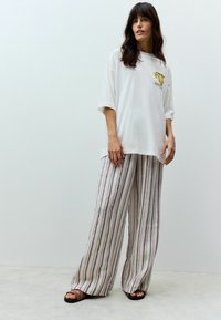 T-shirt bianca oversize con grafica gialla, abbinata a pantaloni a gamba larga a righe in beige e bordeaux. Sandali piatti completano l'outfit.