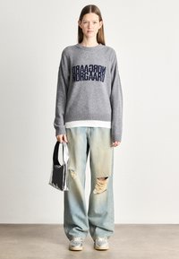 Suéter cinza de malha com texto azul marinho "Norgaard", combinado com jeans desgastados de tom azul claro e uma mala prateada. Calçado casual completa o visual.