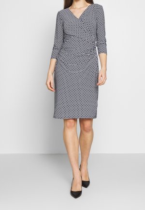 Femme portant une robe portefeuille courte noire et blanche à motifs, manches trois-quarts, et des talons hauts noirs pointus.