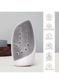 Dispositif de nettoyage facial en silicone, forme courbée, surface texturée avec des nodules arrondis, étanche IPX7, doté de boutons de contrôle, charge magnétique.
