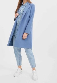 Světle modrý dlouhý kabát z hladké látky, s klopovým límcem a bočními kapsami, nošený přes džínovou košili a světlé džíny.