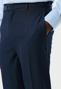 Donkerblauwe, op maat gemaakte broek van gladde stof, voorzien van zijzakken en een platte voorkant met lussen voor een riem.