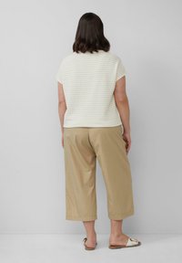 Osoba z ciemnymi włosami do ramion, ubrana w krótki rękaw w paski, beżowe szerokie spodnie 3/4 oraz białe sandały na slip-on.